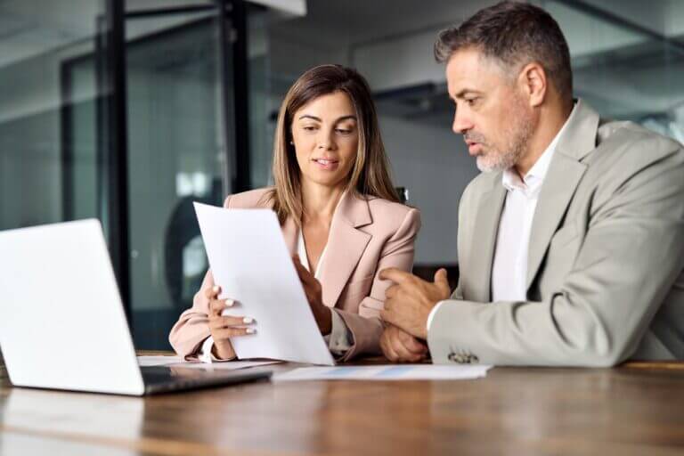 A woman and a man in professional attire are reviewing a document at a wooden desk with a laptop in a modern office.