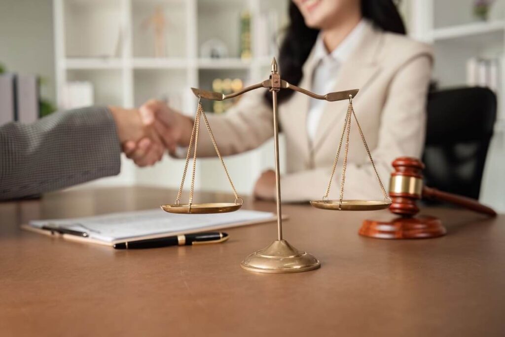 a justice scale, gavel, and documents on a desk, with two people shaking hands in the background