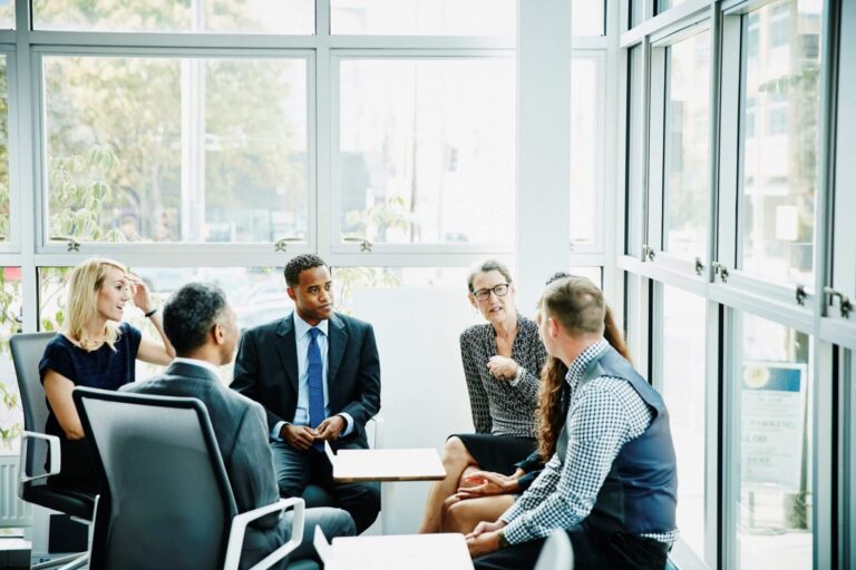 A diverse group of professionals in business attire engage in a discussion in a bright meeting room