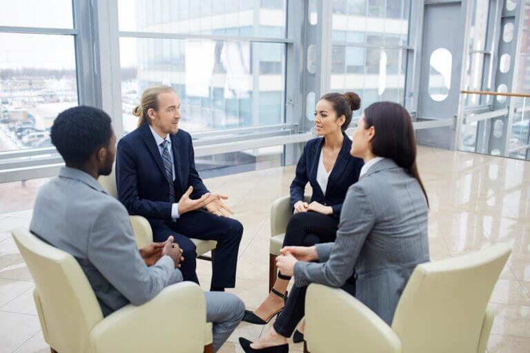 four business professionals in suits are engaged in a discussion, seated in a modern office with large windows
